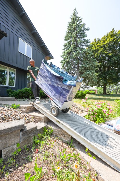 Cottage Grove movers using a ramp over front steps, a Good Stuff Moving crew member wheeling a wrapped sofa down on a dolly